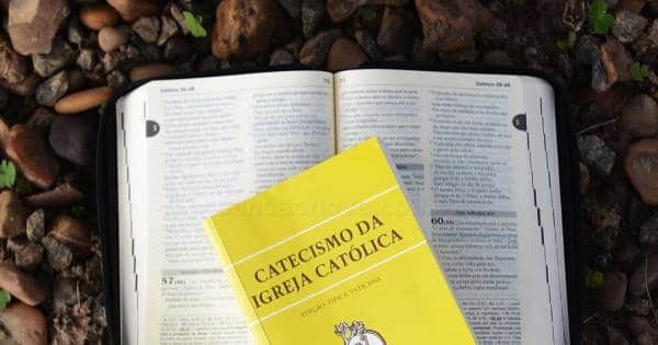 Fundo com algumas pedras de várias cores. Possui uma bíblia aberta e, sobre ela, o livro do catecismo da igreja católica com capa amarela