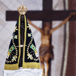 Imagem de Nossa Senhora da Conceição Aparecida com seu manto azul escuro e duas bandeiras pequenas do Brasil simbolizando nossa Padroeira. Ao fundo um crucifixo.